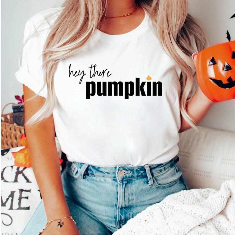 Pode incluir: Camiseta branca com a frase "hey there pumpkin" em escrita preta e letras de bloco, com um pequeno gr&aacute;fico de ab&oacute;bora laranja. A camiseta &eacute; combinada com jeans azuis. Um balde de ab&oacute;bora laranja, uma decora&ccedil;&atilde;o de Halloween, est&aacute; no fundo.