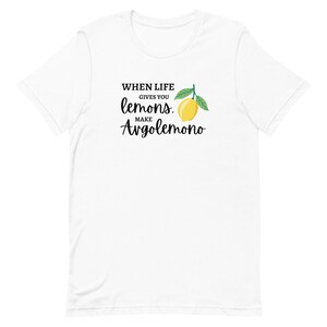 Puede incluir: Una camiseta blanca de manga corta con un gr&aacute;fico de un lim&oacute;n amarillo con hojas verdes y texto en negro que dice: WHEN LIFE GIVES YOU lemons. MAKE Avgolemono. El dise&ntilde;o est&aacute; centrado en la parte del pecho de la prenda.