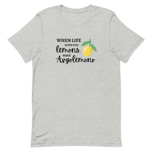Puede incluir: Una camiseta de manga corta en color gris jaspeado claro con un estampado gr&aacute;fico de un lim&oacute;n amarillo con hojas verdes. El texto impreso en negro dice: WHEN LIFE GIVES YOU lemons. MAKE Avgolemono. La prenda tiene cuello redondo y corte.