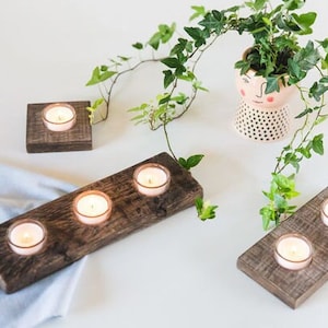 May include: Four wooden candle holders with tea lights, two are rectangular and two are square. The holders are on a white surface with a potted plant with green leaves.