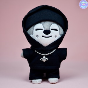 May include: A plush toy wearing a black hoodie with a silver chain necklace and a planet charm. The toy has a gray and white face with a smiling expression.