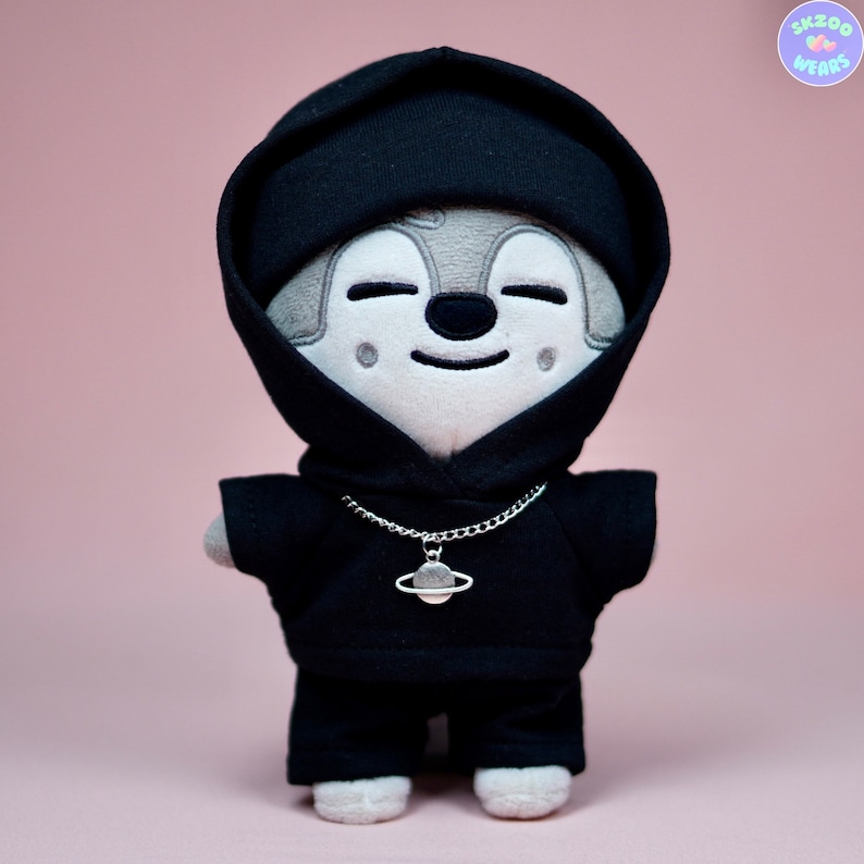 May include: A plush toy wearing a black hoodie with a silver chain necklace and a planet charm. The toy has a gray and white face with a smiling expression.
