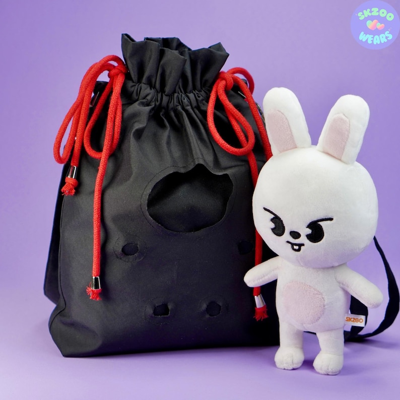 May include: A black drawstring bag with red drawstrings and a white plush bunny with black eyes and a mischievous expression. The bunny is wearing a pink shirt and has the text "SKZOO WEARS" on its back.