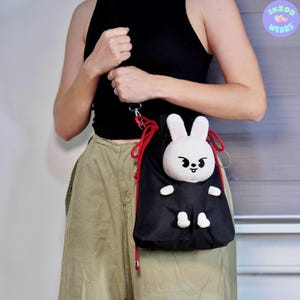 May include: A black drawstring bag with a white bunny plush attached to the front. The bag has a red drawstring and a black strap. The bunny has a black face with a white mouth and black eyes.