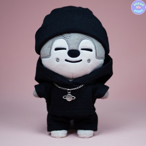 May include: A plush toy wearing a black hoodie, black beanie, and a silver chain with a planet charm. The toy has a gray face with a smiling expression.