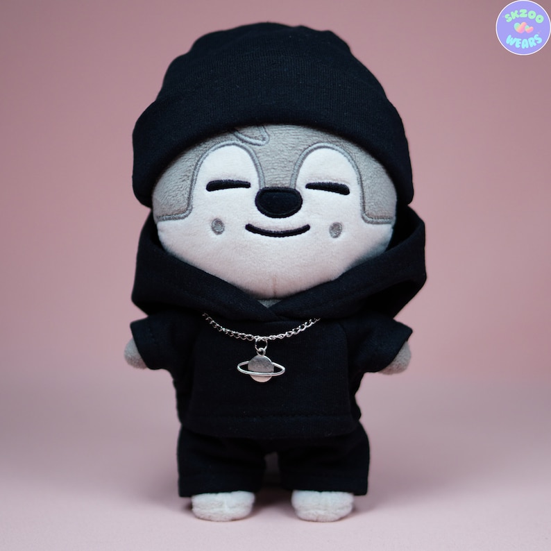 May include: A plush toy wearing a black hoodie, black beanie, and a silver chain with a planet charm. The toy has a gray face with a smiling expression.