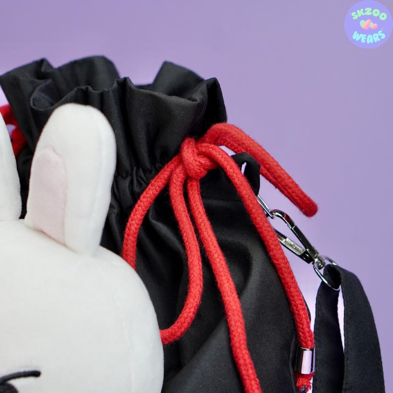 May include: A black drawstring bag with a red drawstring and a white plush bunny attached. The bag has a silver chain strap.