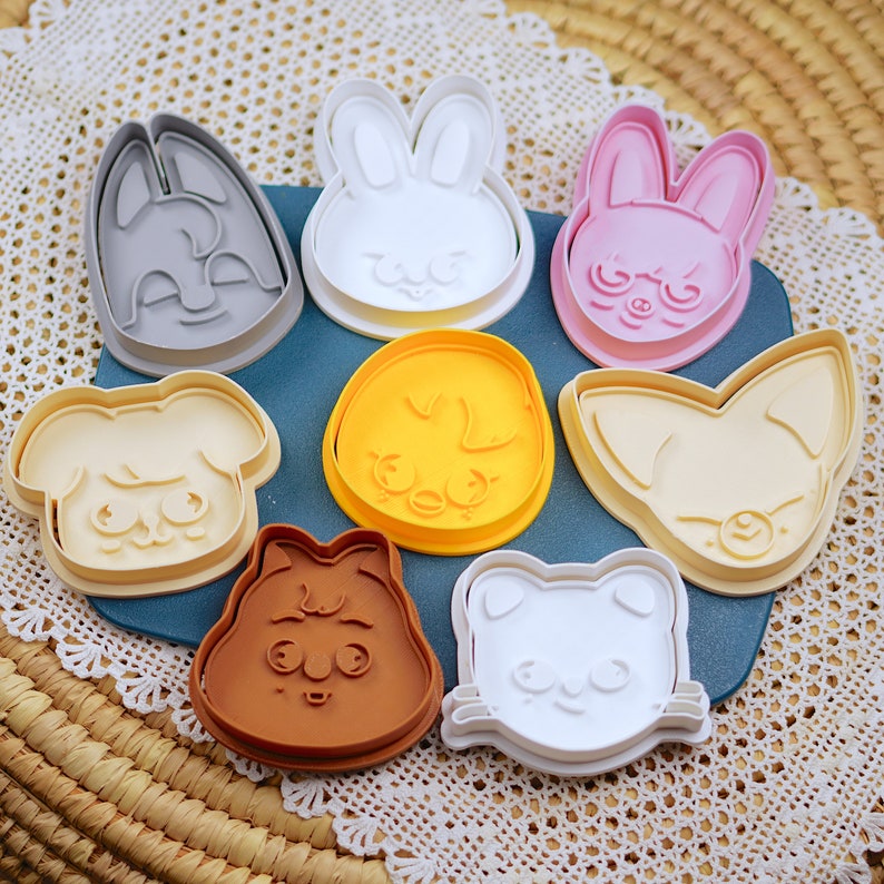 May include: A selection of plastic cookie cutters in animal shapes, such as a rabbit, dog, and cat. The cutters are in shades of grey, white, pink, yellow, and brown, arranged on a blue surface.