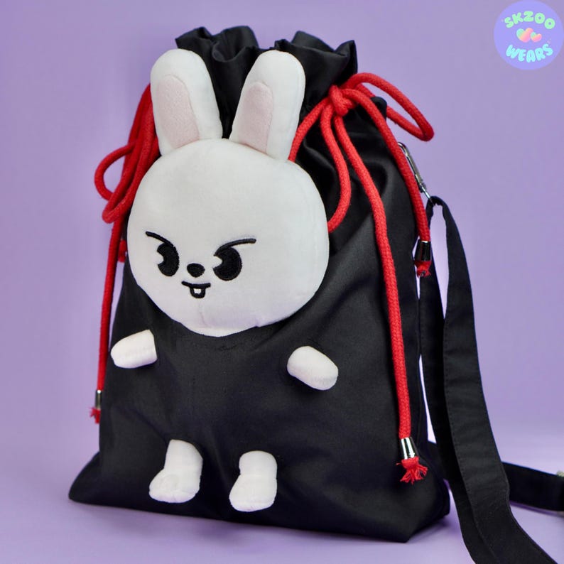 May include: Black drawstring bag with a white plush bunny face attached to the front. The bunny has black eyes and a red mouth. The bag has red drawstrings and a black adjustable strap.