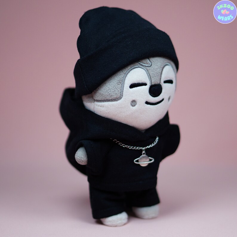 May include: A plush toy wearing a black hoodie, black beanie, and a silver chain with a planet charm. The toy has a gray face with a smiling expression.