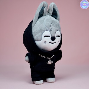 May include: A gray plush toy with a black hoodie and a silver chain necklace with a planet charm. The toy has large ears and a smiling face.