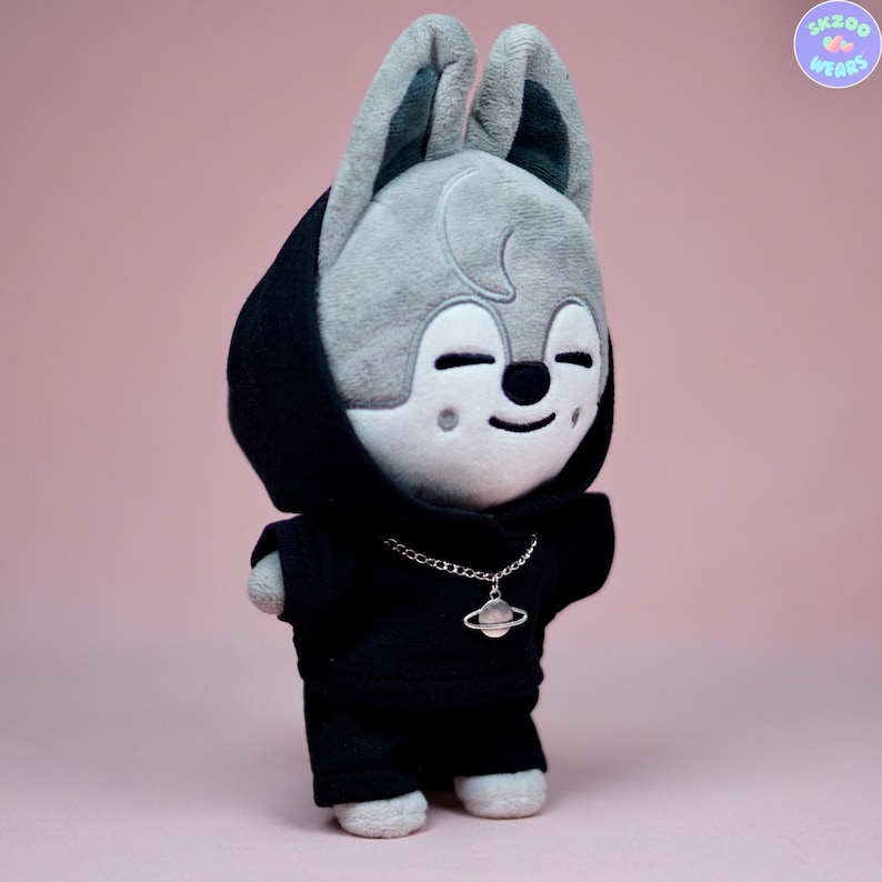 May include: A gray plush toy with a black hoodie and a silver chain necklace with a planet charm. The toy has large ears and a smiling face.