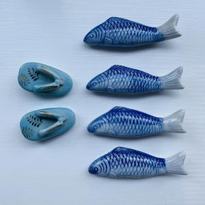 May include: A collection of ceramic fish-shaped chopstick rests and a pair of blue ceramic sandal-shaped rests. The fish are blue with white fins and detailed scale patterns. The sandals are light blue with a gold and black leaf design.