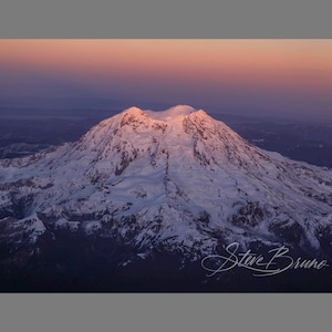 May include: A panoramic view of a snow-capped mountain peak at sunrise, with a pink and purple sky in the background. The mountain is covered in snow and ice, and the surrounding landscape is a mix of forest and hills.