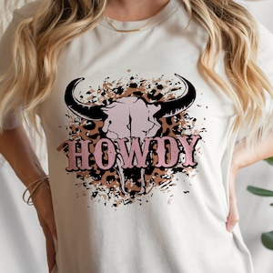 以下が含まれることがあります： 白地にピンクの「HOWDY」のグラフィックがプリントされたTシャツ。黒と白の雄牛の頭蓋骨に、茶色のレオパード柄のアクセントが施されています。
