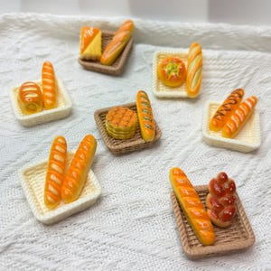 May include: Miniature bread and pastries, including baguettes, rolls, and a braided loaf, are arranged on small woven and knitted trays. The baked goods are painted with realistic details and colors, suggesting a bakery theme.