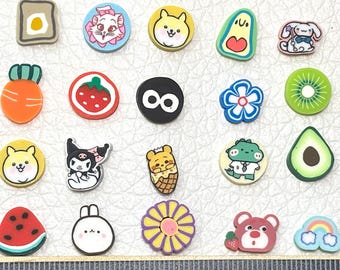 May include: A collection of colorful, cartoon-style charms with various designs, including a fried egg, a cat, a dog, an avocado, a bunny, a carrot, a strawberry, a kiwi slice, a watermelon slice, a flower, a bear, and a rainbow. The charms are arranged on a light-colored surface.