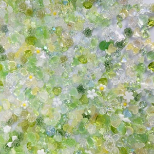May include: A variety of green, clear, and white resin embellishments in the shape of flowers, hearts, and leaves. The embellishments are scattered on a white background.