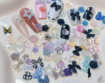 May include: A collection of nail art supplies, including various nail tips with embellishments. The assortment features bows, hearts, stars, butterflies, and flowers in a range of colors, including pink, white, black, and iridescent finishes. The nail art accessories are arranged on a white surface.