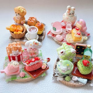 May include: Miniature food and drink figurines, including pastries, cakes, and beverages, arranged on wooden platforms. The collection features a teddy bear, bunny, and other cute characters. The color palette includes pink, yellow, and brown.