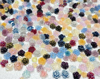 May include: A collection of small, colorful rose-shaped embellishments. The roses are in various shades, including pink, blue, yellow, and purple, with some having a glittery finish. They appear to be made of a translucent material.