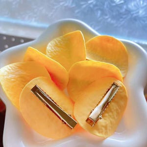 May include: A white, star-shaped bowl filled with several yellow petal-shaped hair clips. Each clip has a gold-colored metal barrette clasp. The clips are arranged in a decorative manner, suggesting a hair accessory product.
