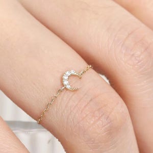 May include: A delicate gold chain ring featuring a crescent moon design. The moon is adorned with small, sparkling gemstones. The ring is worn on a finger, showcasing its elegant and minimalist style.