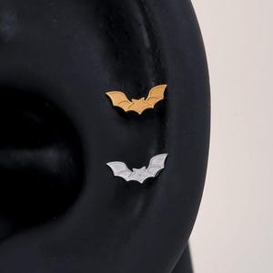Könnte beinhalten: Zwei kleine Ohrstecker in Fledermausform sind an einem dunklen Ohrmodell angebracht. Der obere Ohrstecker ist goldfarben, der untere ist silberfarben. Beide Stecker zeigen eine detaillierte Fledermaussilhouette mit ausgebreiteten Flügeln.