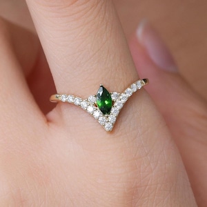 May include: A gold ring with a V-shaped design. The ring features a central, emerald-green gemstone in a marquise cut, surrounded by smaller, clear gemstones. The ring is worn on a finger.