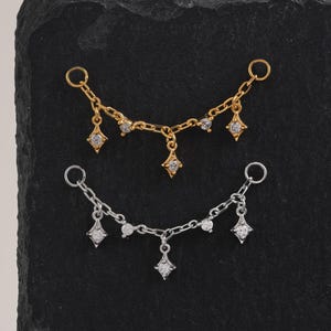 May include: Two delicate chain earrings, one gold-toned and one silver-toned, each adorned with sparkling cubic zirconia. The chains feature small, round stones and dangling diamond-shaped charms. The earrings are displayed on a black surface.