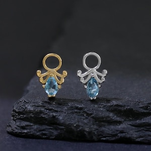 Könnte beinhalten: Zwei Ohrstecker. Einer ist goldfarben, der andere silberfarben. Jeder hat einen tropfenförmigen, hellblauen Edelstein. Die Ohrringe haben ein dekoratives Oberteil-Design. Vor dunklem Hintergrund.