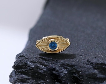 Könnte beinhalten: Ein goldener böser Blick-Anhänger mit einem blauen Edelstein in der Mitte. Der Anhänger liegt auf einer schwarzen Steinoberfläche.