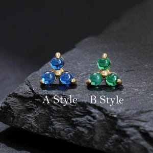 May include: Two pairs of stud earrings, each with a triangular arrangement of three round gemstones. One pair features blue gems, the other green. The earrings are on a dark, textured surface. Text on the surface reads "A Style" and "B Style".