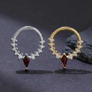 May include: Two hoop earrings, one silver and one gold, each featuring a row of small, clear stones. A red, diamond-shaped stone hangs from the bottom of each earring. The earrings are displayed on a dark gray surface.
