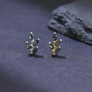 May include: Two leaf-shaped stud earrings. One earring is silver-toned, the other is gold-toned. Each earring features small, dark green gemstones arranged to resemble leaves. The earrings are displayed on a dark surface.