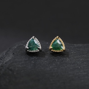 Puede incluir: Un par de pendientes de botón con piedras preciosas. Un pendiente tiene un engaste plateado con una piedra triangular verde, y el otro tiene un engaste dorado con una piedra verde similar. Los pendientes se muestran sobre una superficie oscura y texturizada.