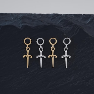 May include: Four small sword-shaped earrings in gold and silver tones are displayed on a dark, textured surface. The earrings have a circular top and a dagger-like design, showcasing a minimalist aesthetic.