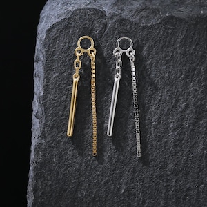 Può includere: Un paio di orecchini pendenti, uno dorato e uno argentato, esposti su una superficie scura e strutturata. L'orecchino dorato presenta un pendente a barra piena e una catena, mentre l'orecchino argentato ha un design simile.