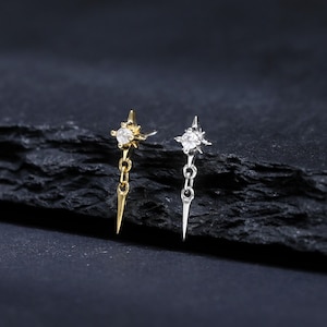 May include: Two stud earrings, one gold-toned and one silver-toned, each featuring a four-pointed star design with a central clear stone and a small dangling spike element attached by a short chain link, presented as a pair of delicate jewellery.