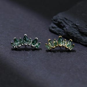 Könnte beinhalten: Zwei Paar Ohrringe, jeweils mit einer Anordnung grüner Edelsteine in Blattform. Ein Paar hat eine silberfarbene Fassung, das andere eine goldfarbene Fassung. Die Ohrringe werden auf einer dunklen Oberfläche präsentiert.