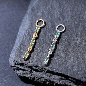 Op de afbeelding: Twee verticale bedels, één in goudkleur en één in zilverkleur, elk voorzien van drie kleine rechthoekige groene stenen in een lineaire opstelling met een ronde ring aan de bovenkant voor bevestiging aan een ketting of oorbel.