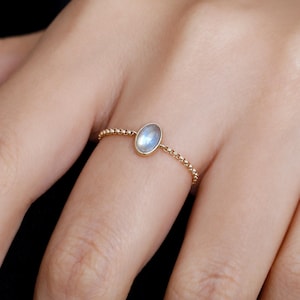 Puede incluir: Un anillo de oro con una pequeña piedra preciosa ovalada de color azul claro. La banda del anillo está adornada con pequeñas cuentas doradas espaciadas uniformemente. El anillo se muestra en un dedo, destacando su delicado diseño.