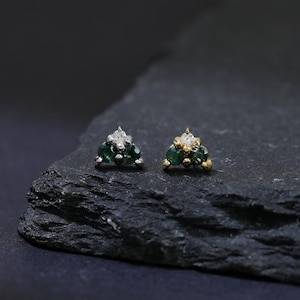 May include: Two small stud earrings with a triangular design. Each earring features a cluster of green gemstones and small white gemstones. One earring has a silver setting, and the other has a gold setting. The earrings are displayed on a dark gray, textured surface.