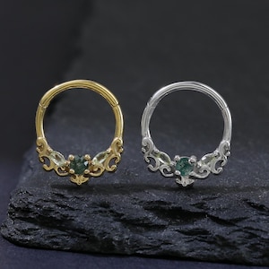 Könnte beinhalten: Zwei Nasenringe, einer goldfarben und einer silberfarben, werden gezeigt. Jeder Ring hat ein dekoratives Design mit einem kleinen, grünen Edelstein in der Mitte. Die Ringe sind vor einem dunklen, strukturierten Hintergrund platziert.