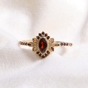 May include: A gold ring featuring a central, oval-cut, deep red gemstone. The ring is embellished with smaller, reddish-brown stones around the main gem and along the band. The ring is displayed on a soft, white fabric background.