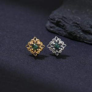 Op de afbeelding: Twee oorstekers met een bloemmotief. Eén oorbel is goudkleurig, de andere zilverkleurig. Beide hebben een donkergroene middensteen en staan tegen een donkere achtergrond.