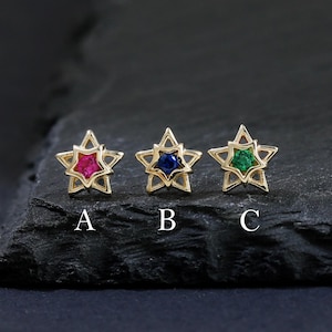 May include: Three gold star-shaped stud earrings with red, blue, and green gemstone centres. Each earring is labelled A, B, and C. The earrings are displayed on a dark, textured surface. The background is dark.