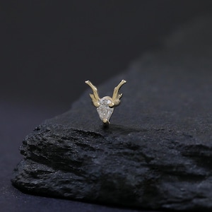 Puede incluir: Un pendiente dorado con una piedra preciosa clara en forma de pera en el centro. El pendiente presenta un diseño de cuerno estilizado. La joya se exhibe sobre una superficie oscura y texturizada sobre un fondo negro.