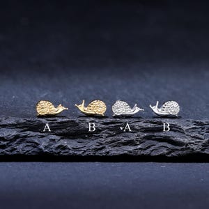 Puede incluir: Cuatro pares de pendientes de botón con forma de caracol. Dos pares son dorados y dos plateados con pequeñas piedras brillantes. Cada par está etiquetado con una letra, A o B. Los pendientes se muestran sobre una superficie oscura y texturizada.