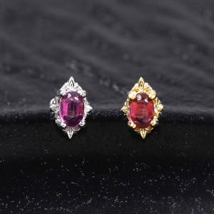 Pode incluir: Dois brincos de pino com pedras preciosas rosa-escuras de corte oval em armações de metal decorativas. Um brinco tem um acabamento prateado, enquanto o outro é dourado. Ambas as peças exibem um detalhe metálico pontiagudo em forma de estrela.